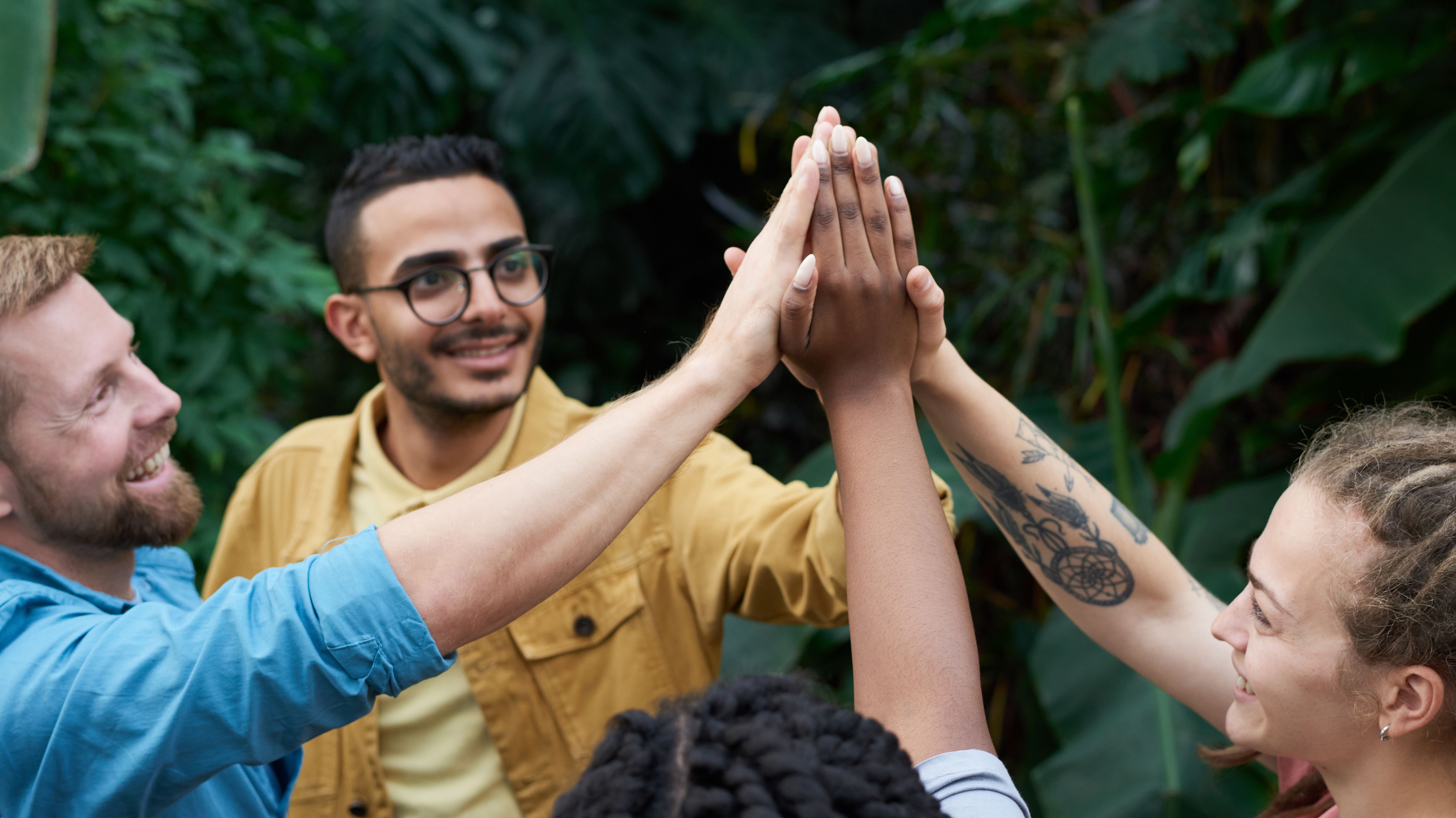 Four colleagues join hands in a high-five surrounded by greenery.